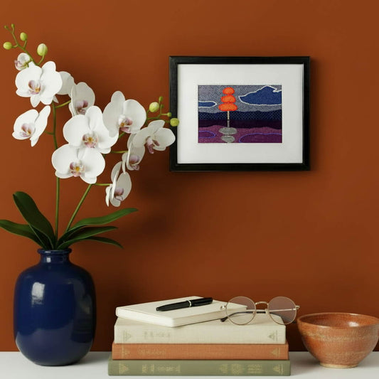 Framed fabric artwork hanging on an orange wall with a modern console table, vase, books, and notebook with pen