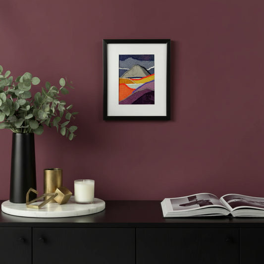 Framed fabric artwork hanging on a purple wall with a console table, book, and plant in a contemporary home