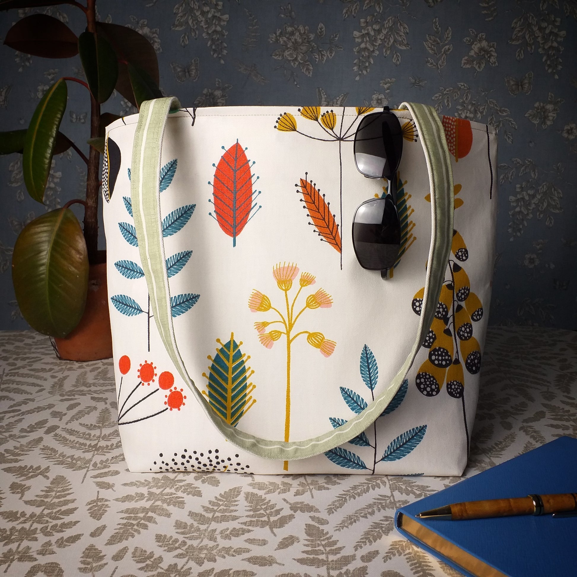 Large tote bag with multi-colour wild flowers design on off-white background sitting on a table with a notebook, pen, and plant