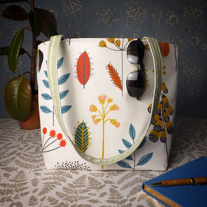 Large tote bag with multi-colour wild flowers design on off-white background sitting on a table with a notebook, pen, and plant