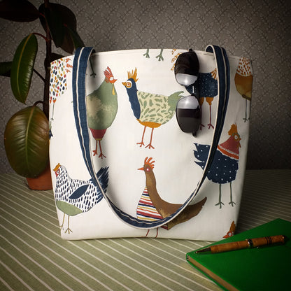 Large tote bag with multi-colour hens design on beige background sitting on a table with a notebook, pen, and plant