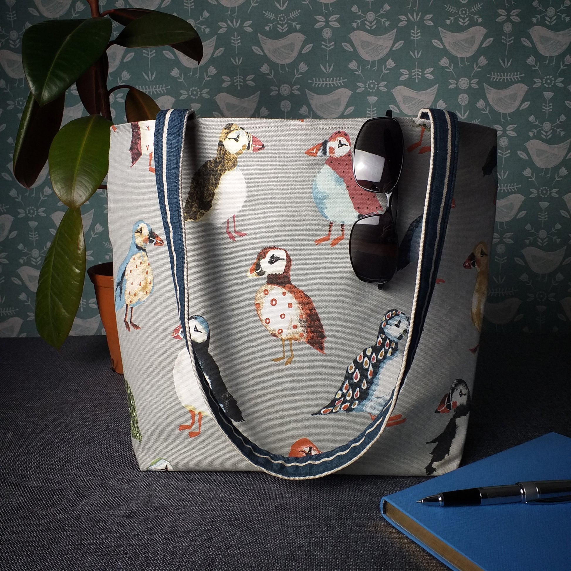 Large tote bag with multi-colour puffins design on grey background sitting on a table with a notebook, pen, and plant