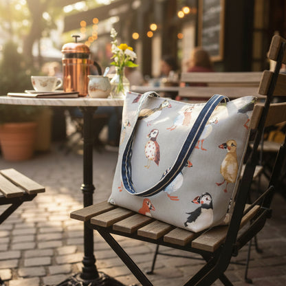 Large tote bag with multi-colour puffins design on grey background sitting on an outdoor chair at a boutique cafe