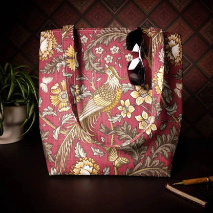 Large tote bag with pheasants and flowers design on red background sitting on a table with a notebook, pen, and plant
