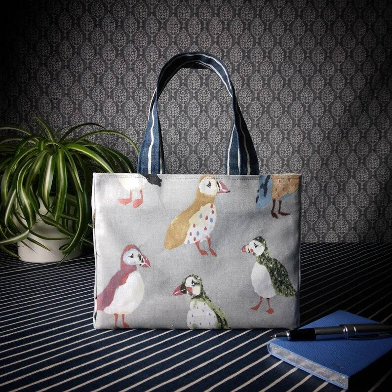 Mini tote bag with multi-colour puffins design on grey background sitting on a table with notepad, pen and plant