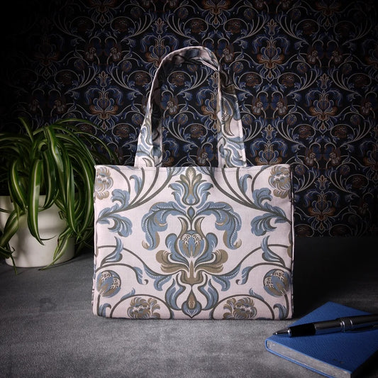Mini tote bag with grey and brown baroque style floral design on taupe background sitting on a table with notepad, pen and plant
