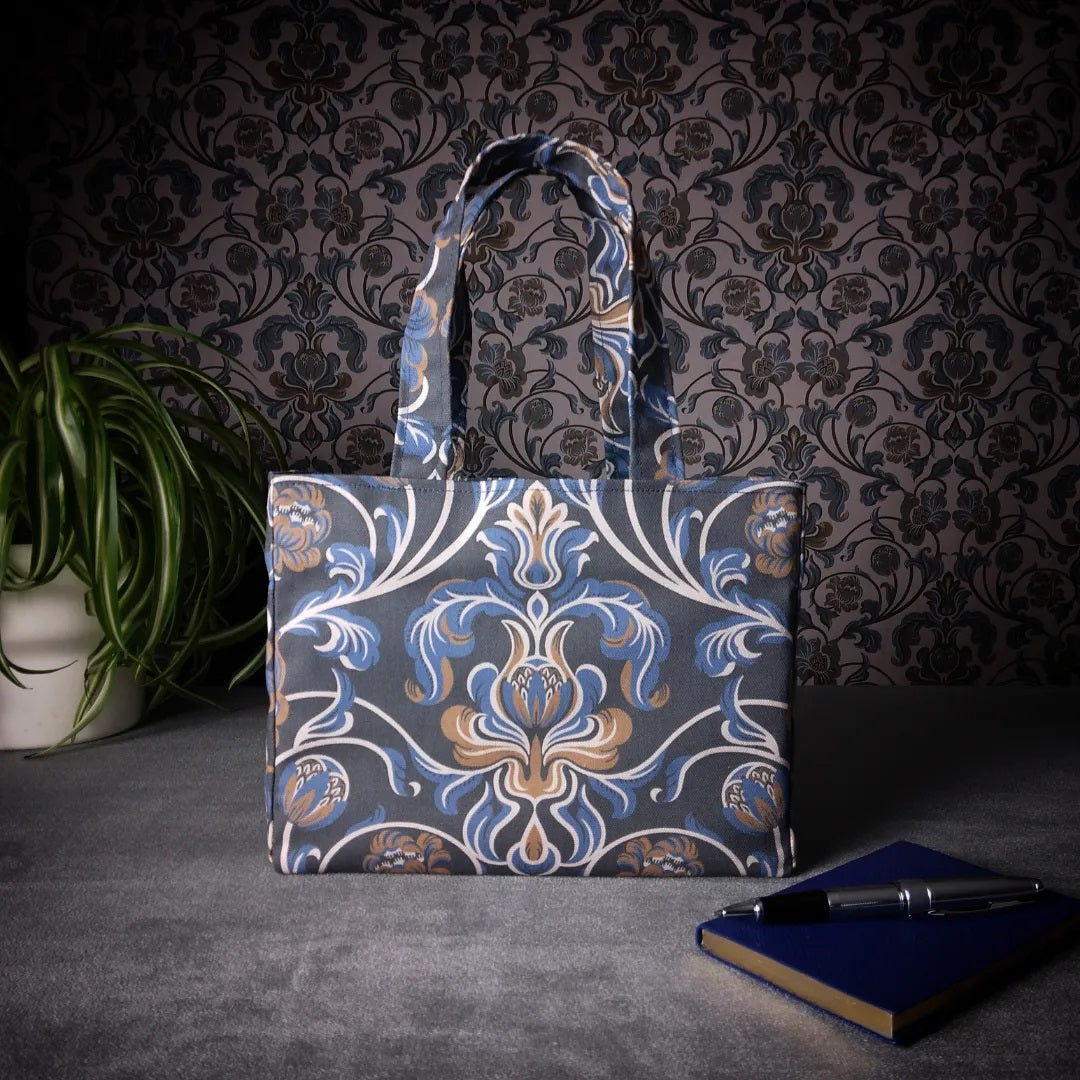 Mini tote bag with white, brown, and blue baroque style floral design on navy background sitting on a table with a notebook, pen, and plant