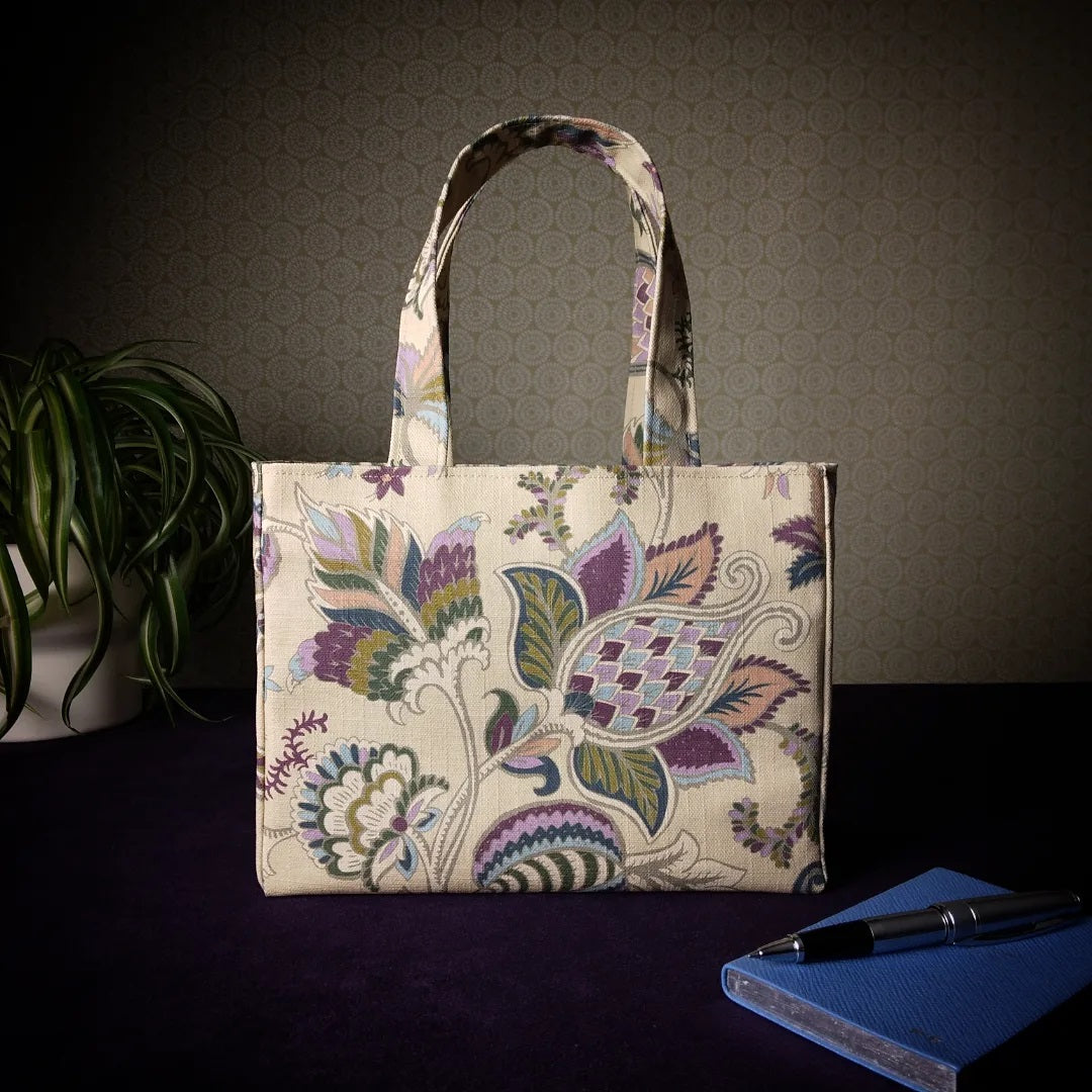 Mini tote bag with multi-colour floral print on beige background sitting on a table with notepad, pen and plant