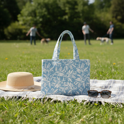 Mini tote bag with white wildflower silhouette design on light blue background sitting on a picnic blanket in a community park