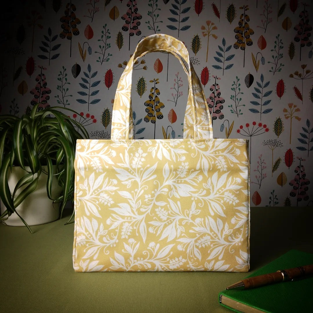 Mini tote bag with white wildflower silhouette design on yellow background sitting on a table with notepad, pen and plant