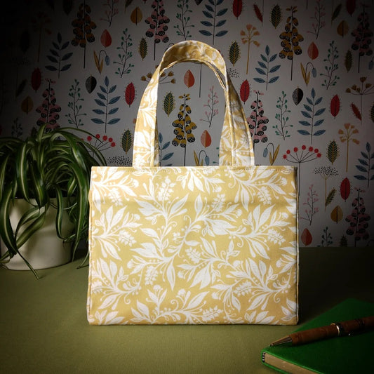 Mini tote bag with white wildflower silhouette design on yellow background sitting on a table with notepad, pen and plant