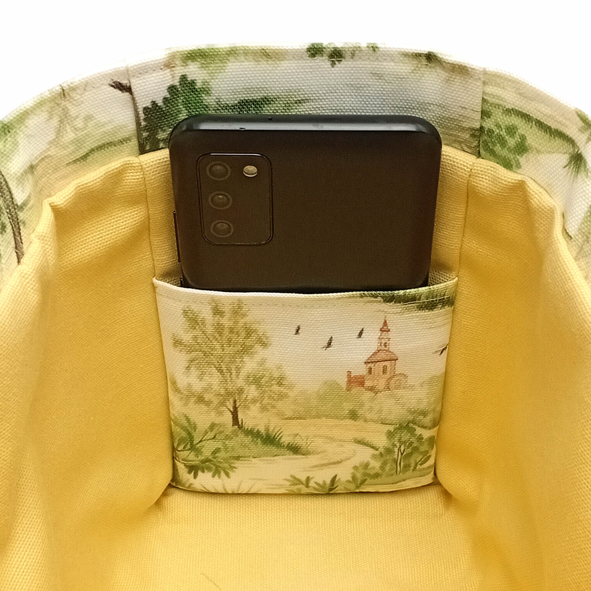 A close-up of a side slip pocket holding a black smartphone inside a handmade mini cotton tote bag with pale yellow lining and a green toile exterior.