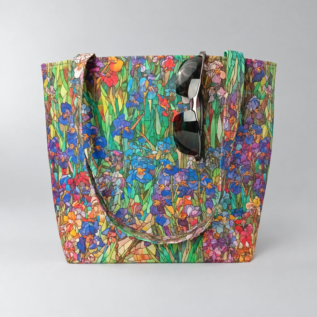 A large handmade cotton tote bag featuring a colourful floral and iris pattern with green stems and blue, purple, pink, and red blossoms. The bag has two shoulder straps and is shown with a pair of black sunglasses.