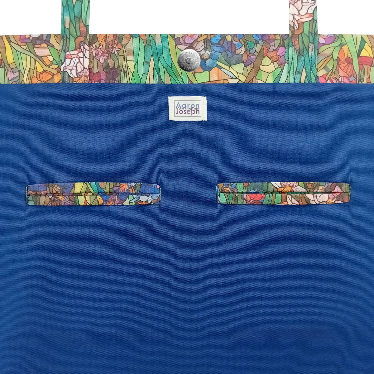 A handmade large cotton tote bag blue interior with a colourful stained-glass style iris pattern trim along the top edge, top handles, and two double welt pockets.