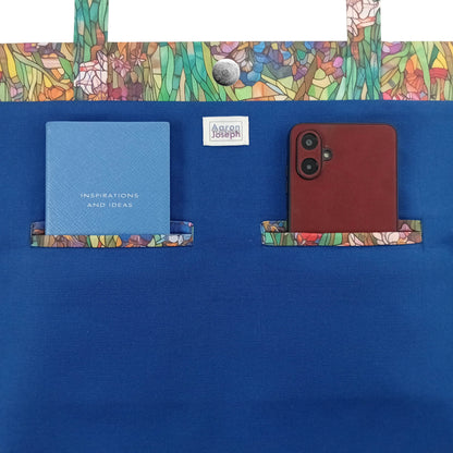 A handmade large cotton tote bag blue interior with a colourful stained-glass style iris pattern trim along the top edge, top handles, and a notebook and mobile phone inside two double welt pockets.