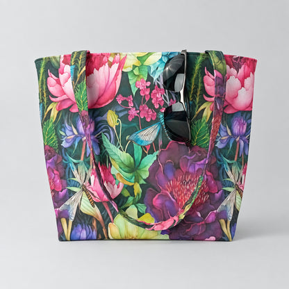 A large handmade cotton tote bag featuring a colourful botanical print with pink flowers, green leaves, and dragonflies, shown against a neutral light grey background with sunglasses hanging from the top of the tote bag.