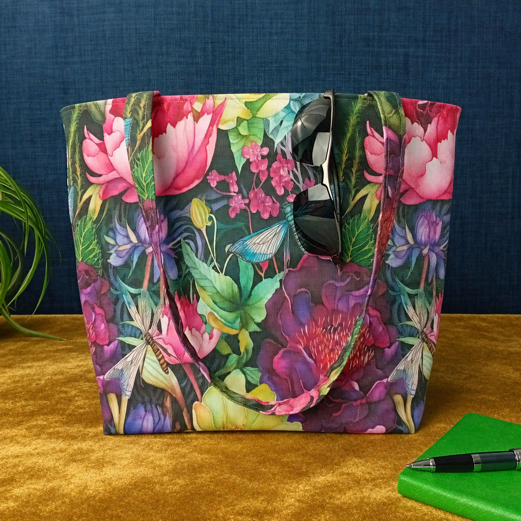 A large handmade cotton tote bag with a vibrant, multicoloured floral and botanical print sits on a mustard-yellow velvet surface against a dark blue textured background. A pair of black sunglasses rests on the bag, and a green notebook with a pen is visible in the bottom right corner.