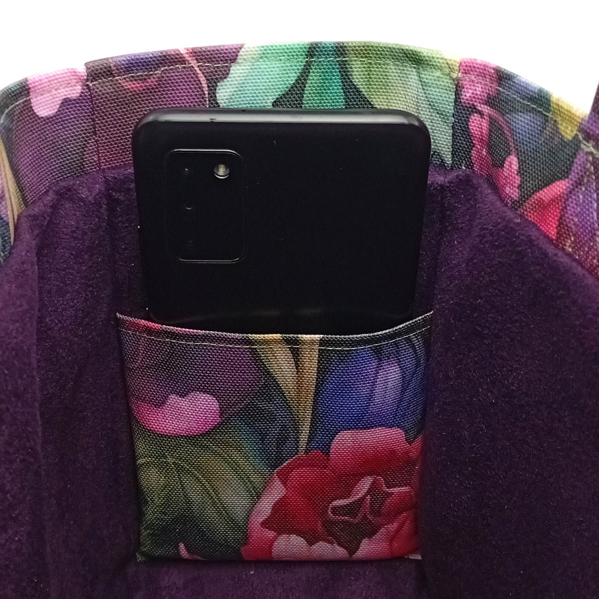 A close up of a handmade mini cotton tote bag interior with a dark purple velvet lining featuring a floral print slip pocket with black smartphone, and floral print trim.