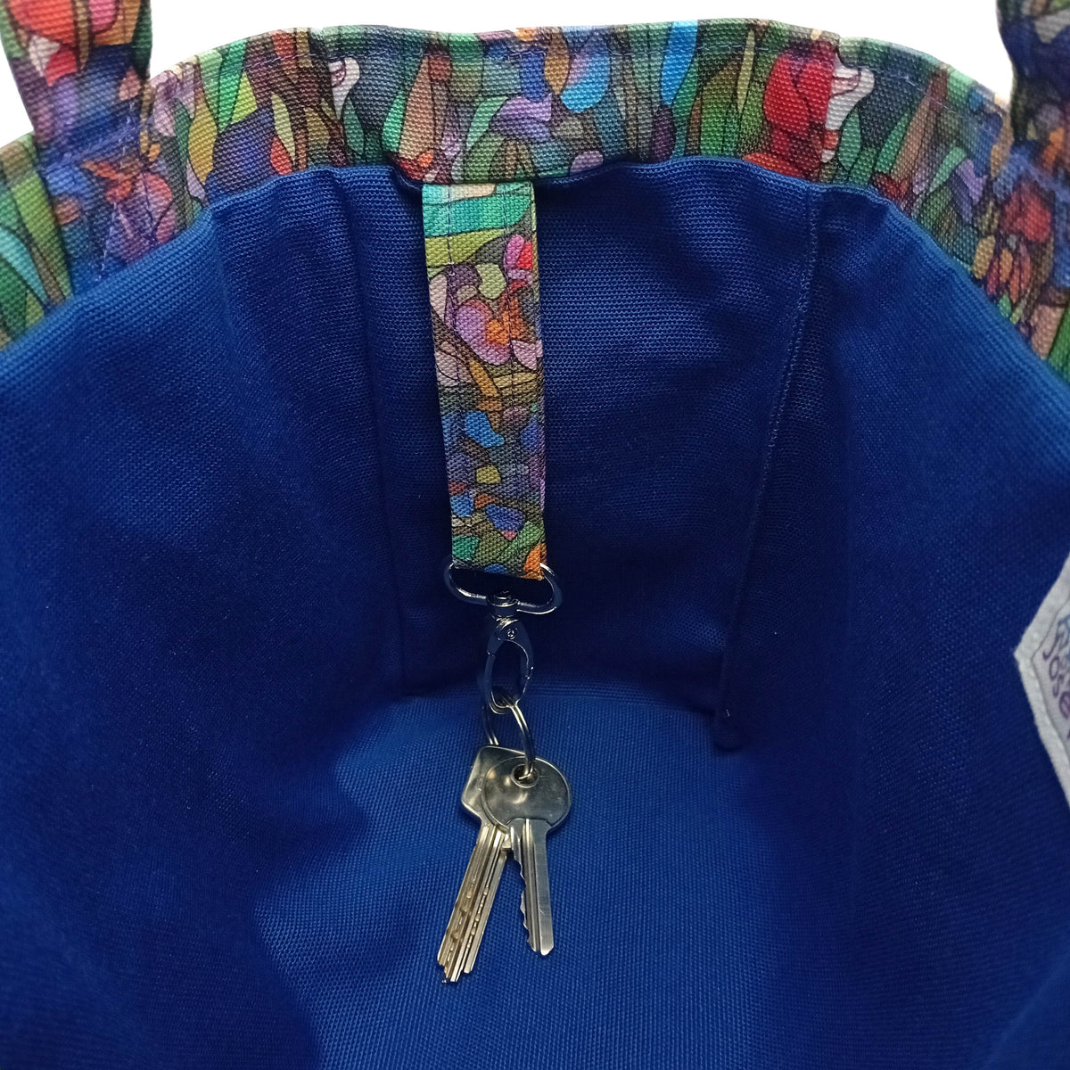Close-up of a handmade mini cotton tote bag interior with a blue interior and colourful floral-patterned strap with key clasp holding two metal keys.