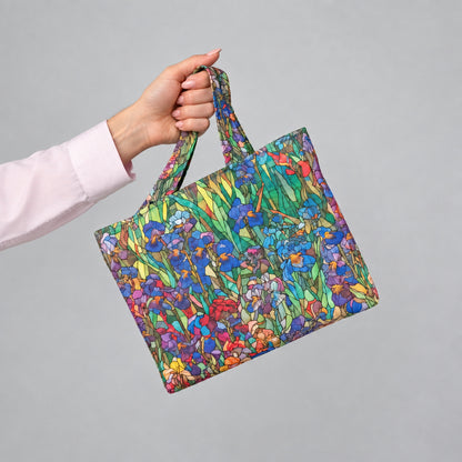 A hand holding a handmade mini cotton tote bag with a colourful floral print featuring blue and purple irises, red, and yellow flowers with green leaves, set against a plain light grey background.