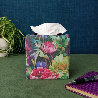 A handmade square cotton tissue box cover with a colourful floral and dragonfly print sits on a green tabletop against a blue textured wall. A white tissue is pulled from the top, with a green plant and a purple notebook visible in the background.