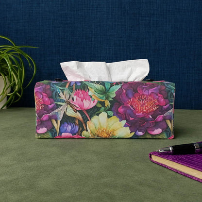 A handmade rectangular cotton tissue box cover with a colourful floral and dragonfly print with a plant and a purple notebook in the background.