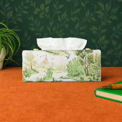 A handmade rectangular cotton tissue box cover with a printed landscape scene in soft green, beige, and brown tones on an off-white background, with a plant on the left and a green notebook with a pen on the right.