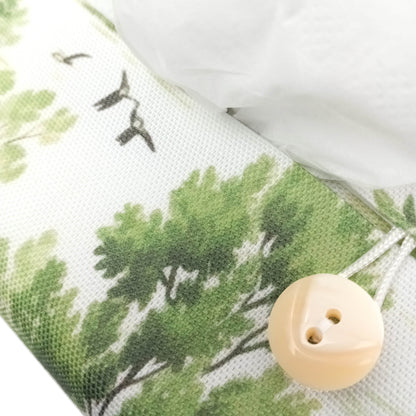 A close-up of a handmade rectangular cotton tissue box cover with a green tree and bird print on an off-white background, featuring a cream-colored button closure.