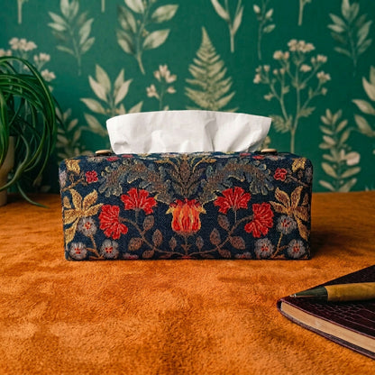 A handmade polycotton rectangular tissue box cover with a dark navy blue background and an ornate floral pattern in red, gold, and beige. It sits on a tabletop with a blue textured wall behind it, a green plant on the left, and a red notebook with a pen on the right.