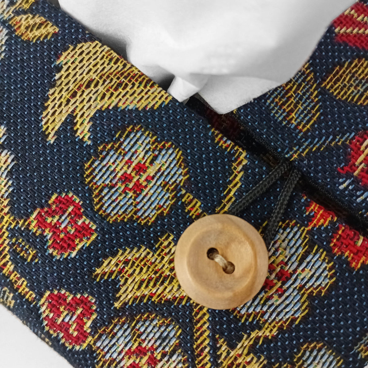 Close-up of a rectangular tapestry fabric tissue box cover with a dark navy base, woven floral motifs in gold, red, and light blue, and a small round natural wood button.