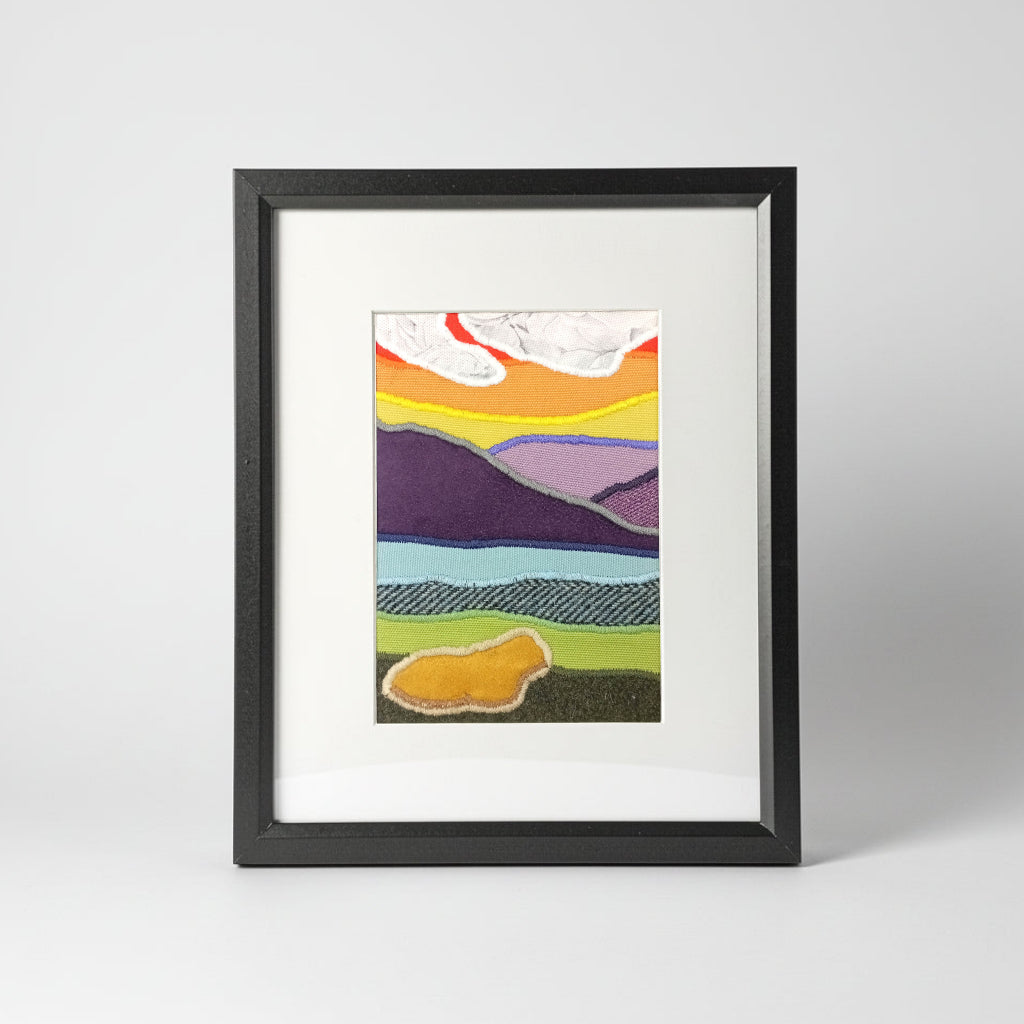 A vertical framed handmade abstract landscape fabric artwork with colourful layered hills, a sunrise with white clouds, and a highland cow presented in a black frame with a white mat.