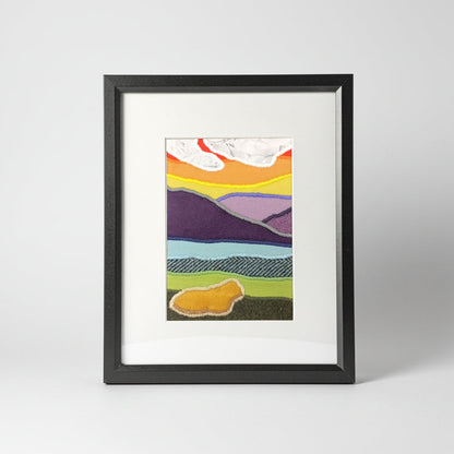 A vertical framed handmade abstract landscape fabric artwork with colourful layered hills, a sunrise with white clouds, and a highland cow presented in a black frame with a white mat.