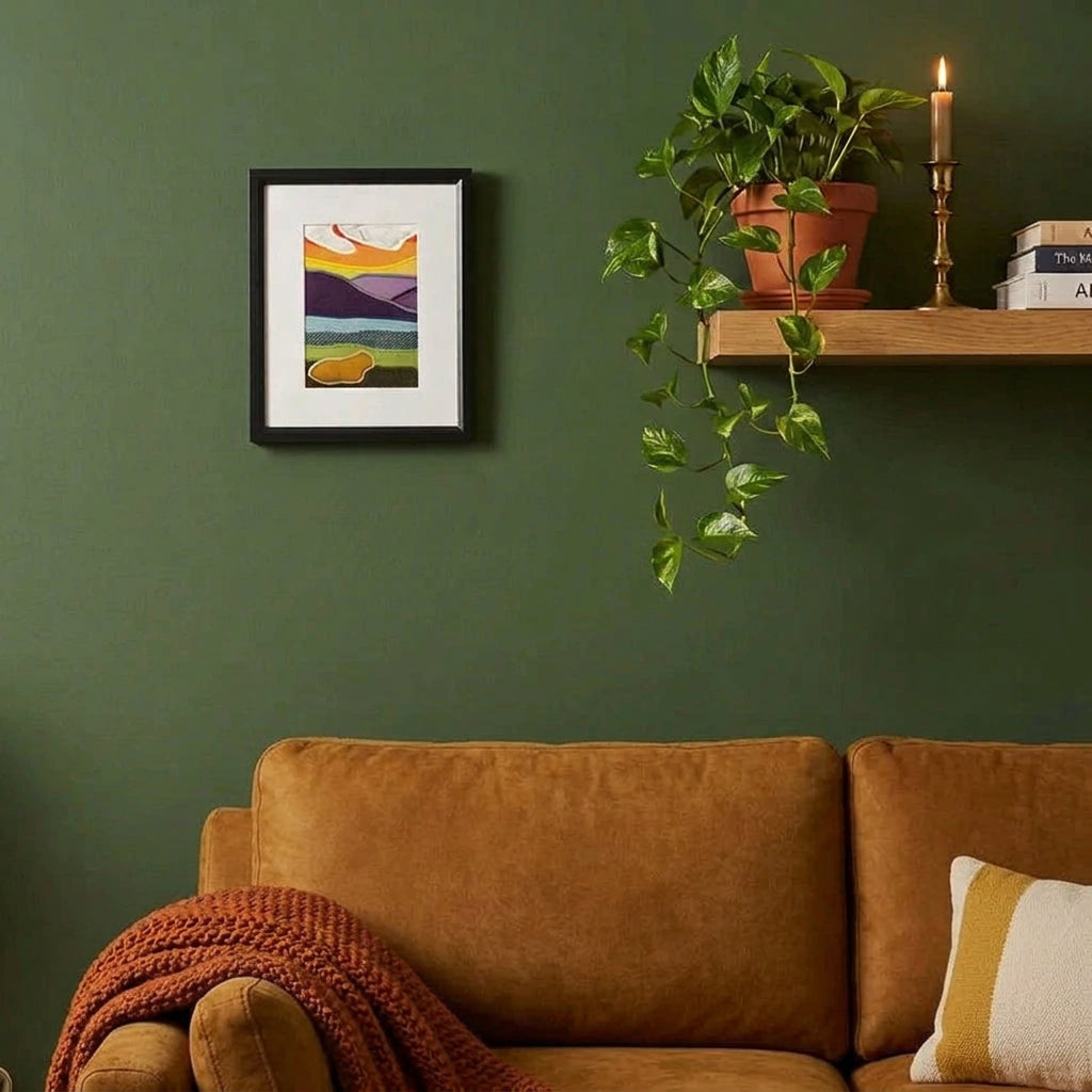 A framed handmade fabric abstract landscape art print on a living room wall with a shelf, lamp. plant, and armchair.
