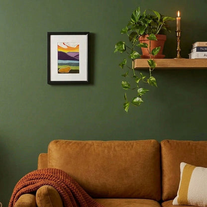 A framed handmade fabric abstract landscape art print on a living room wall with a shelf, lamp. plant, and armchair.