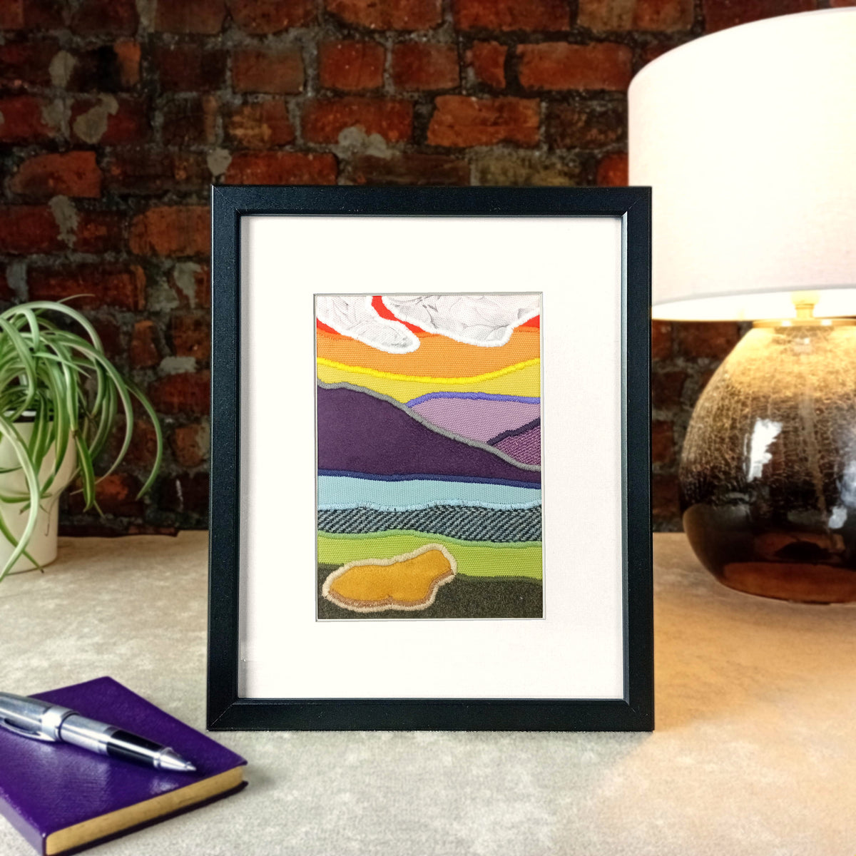A vertical framed handmade abstract fabric landscape collage artwork with colourful layered hills, clouds, and a stylised highland cow, displayed on a tabletop next to a purple notebook, pen, plant.