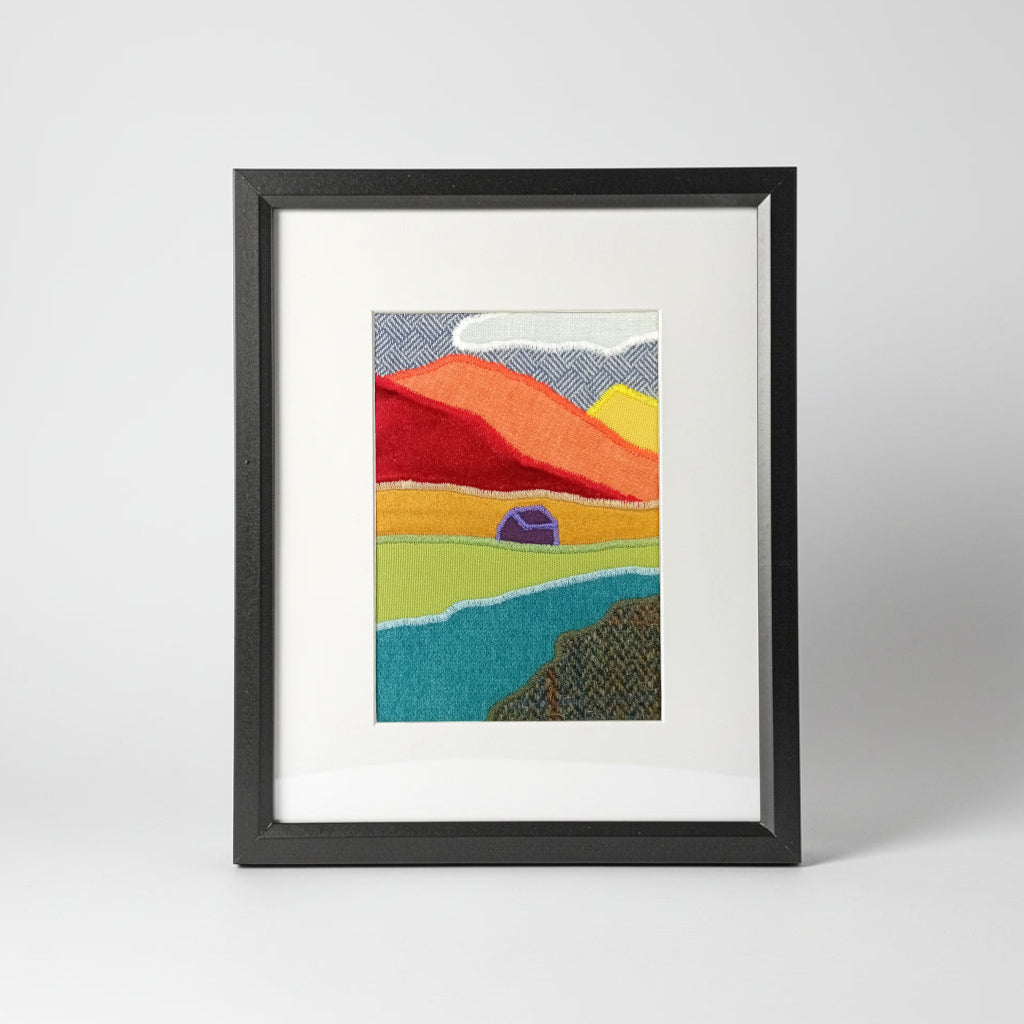 A vertical framed handmade abstract fabric landscape artwork featuring textured, patchwork-like mountains, river, and house, presented in a black frame with a white mat
