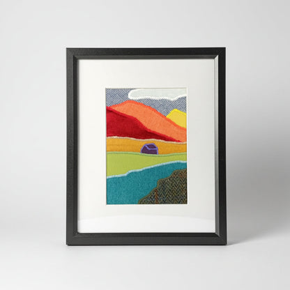 A vertical framed handmade abstract fabric landscape artwork featuring textured, patchwork-like mountains, river, and house, presented in a black frame with a white mat