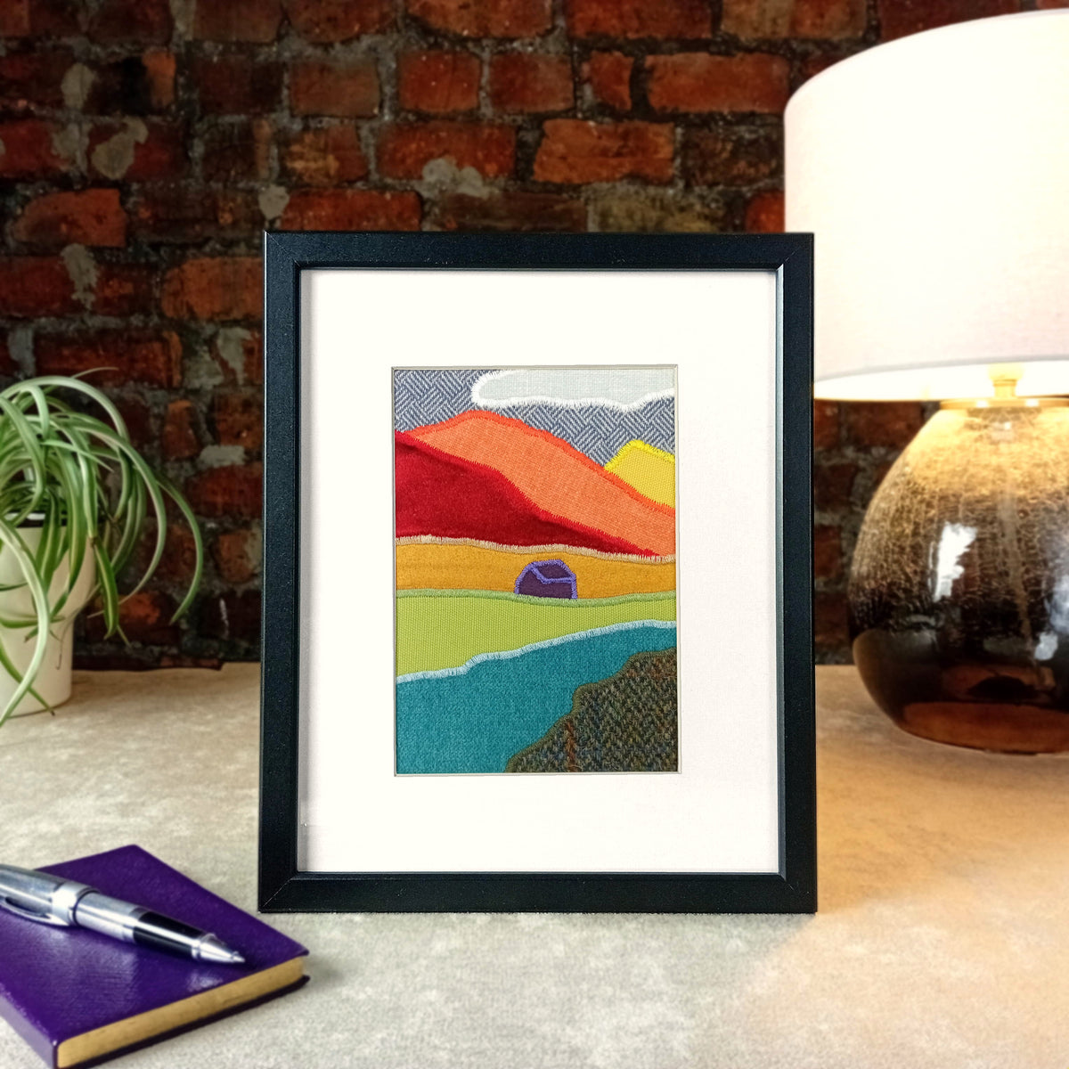 A vertical framed handmade abstract fabric landscape artwork displayed on a tabletop next to a plant, notebook, and lamp against a brick wall background.
