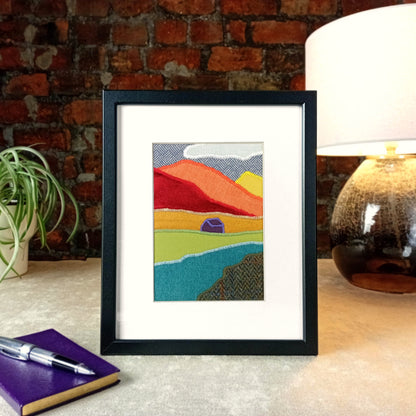 A vertical framed handmade abstract fabric landscape artwork displayed on a tabletop next to a plant, notebook, and lamp against a brick wall background.
