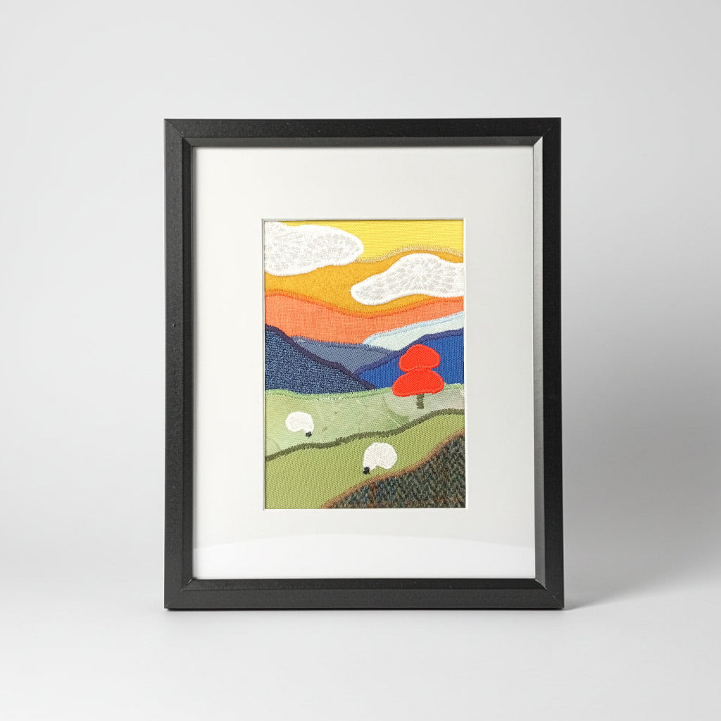A vertical framed collage style fabric artwork featuring a stylized landscape with rolling green hills, white sheep, an orange tree, blue mountains, and a yellow-orange sky, presented in a black frame with a white mat.