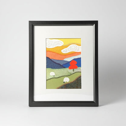 A vertical framed collage style fabric artwork featuring a stylized landscape with rolling green hills, white sheep, an orange tree, blue mountains, and a yellow-orange sky, presented in a black frame with a white mat.