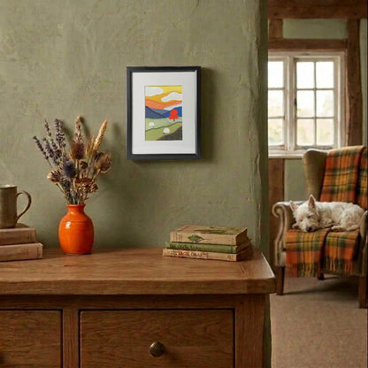 A vertical framed handmade fabric artwork featuring a stylised landscape with rolling green hills, white sheep, and blue mountains hanging on a cottage wall.