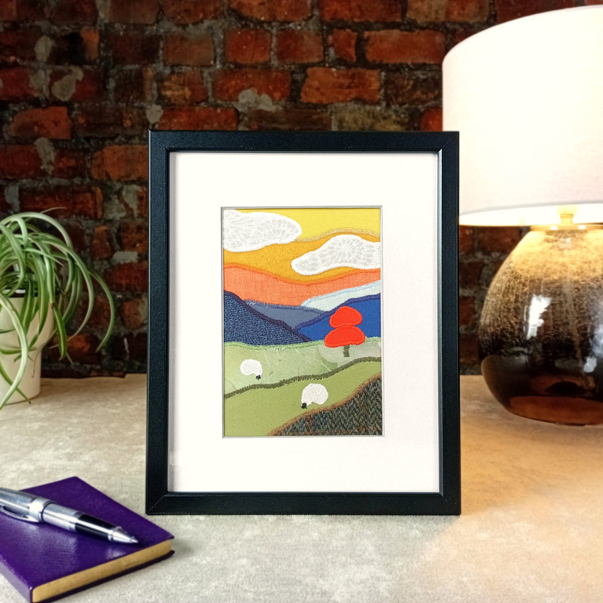 A vertical framed handmade fabric artwork depicting a stylized landscape with rolling green hills, white sheep, a red tree, and layered mountains under a yellow and orange sky, displayed on a tabletop with a brick wall, lamp, notebook, and pen in the background.