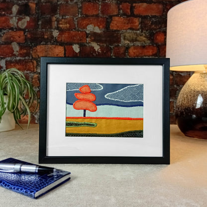 Framed fabric artwork sitting on a table with a lamp and plant in front of a brick wall