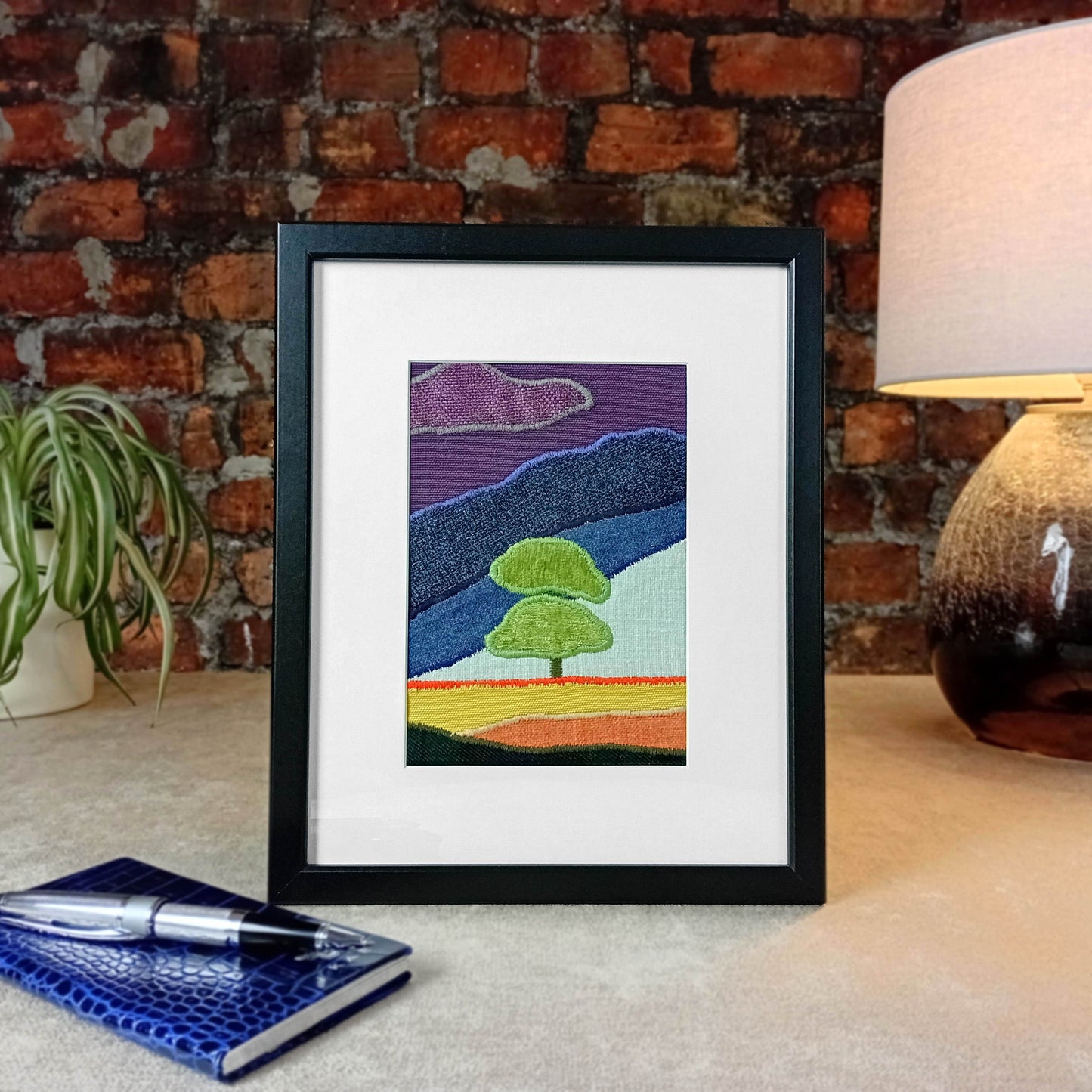 Framed fabric artwork sitting on a table with a lamp and plant in from of a brick wall