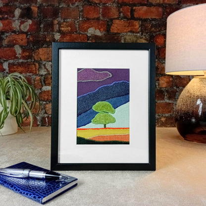 Framed fabric artwork sitting on a table with a lamp and plant in from of a brick wall