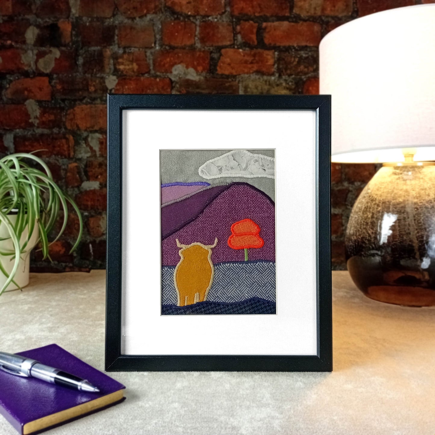 Framed fabric art sitting on a table with lamp and plant in front of a brick wall