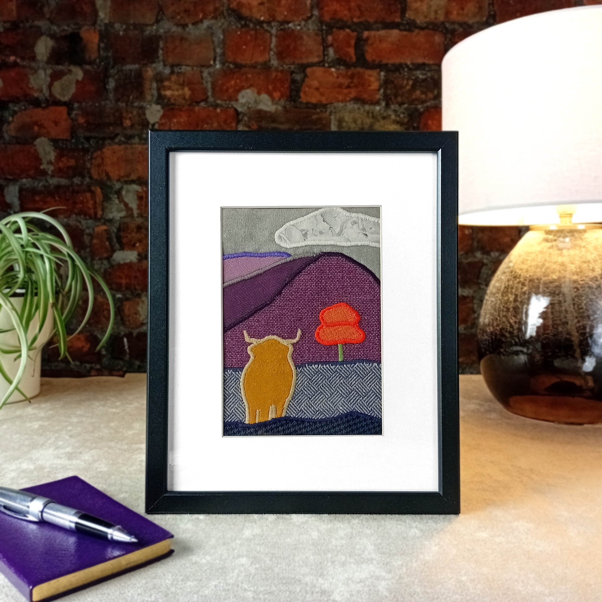 Framed fabric art sitting on a table with lamp and plant in front of a brick wall