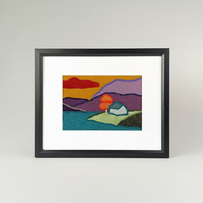 Framed fabric landscape scene of a cottage by the lake with a tree and mountains