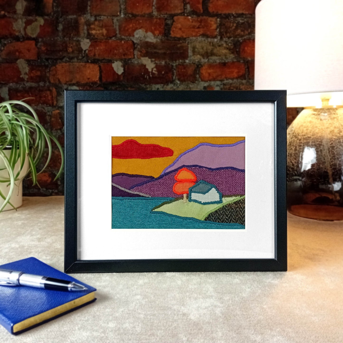 Framed fabric landscape scene of a cottage by the lake with a tree and mountains sitting on a table with a lamp, plant in front of a brick wall